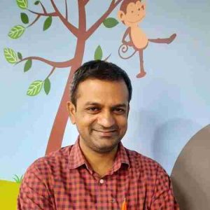 Dr vallikiran - Pathway Hospital Smiling man in a red plaid shirt posing in front of a mural showing a tree and a cartoon monkey.