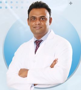 dr-umesh-n-plastic-surgeon-bangalore 2 - Pathway Hospital Male doctor in a white coat with a maroon polka-dot tie, arms crossed, posing for a professional portrait against a light blue background with circular design.