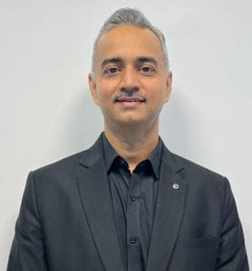 Dr ambrish sharma - Pathway Hospital Head-and-shoulders portrait of a man in a black suit and black shirt against a light gray background, in a formal business pose.